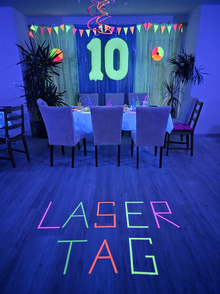 Laser Tag Party zuhause mit Neon-Dekoration, Schwarzlicht und großem LASER TAG-Schriftzug im festlich dekorierten Partyraum.
